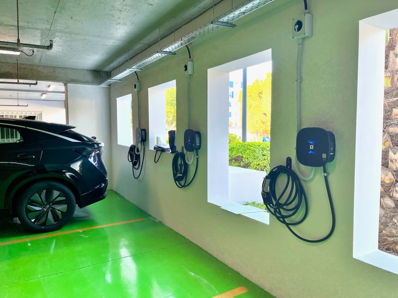 A row of wall-mounted AC chargers with tethered cables along the green-marked charging bays in the parking garage